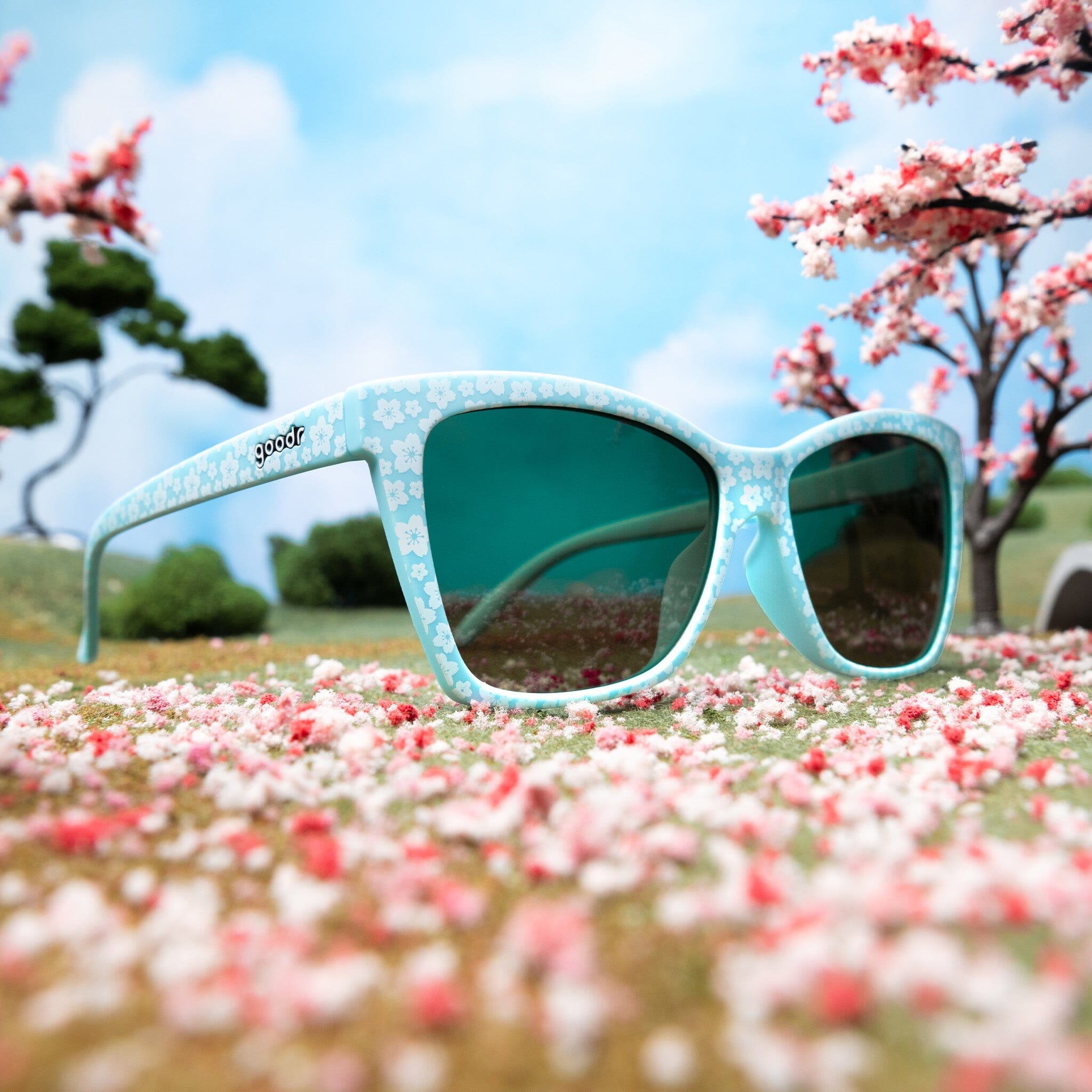 Three-quarter angle view of light blue cherry blossom sunglasses sitting in a miniature landscape of cherry blossom trees.