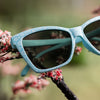 Light blue cherry blossom sunglasses sitting in a mini landscape of cherry blossom trees with a blurred green background.